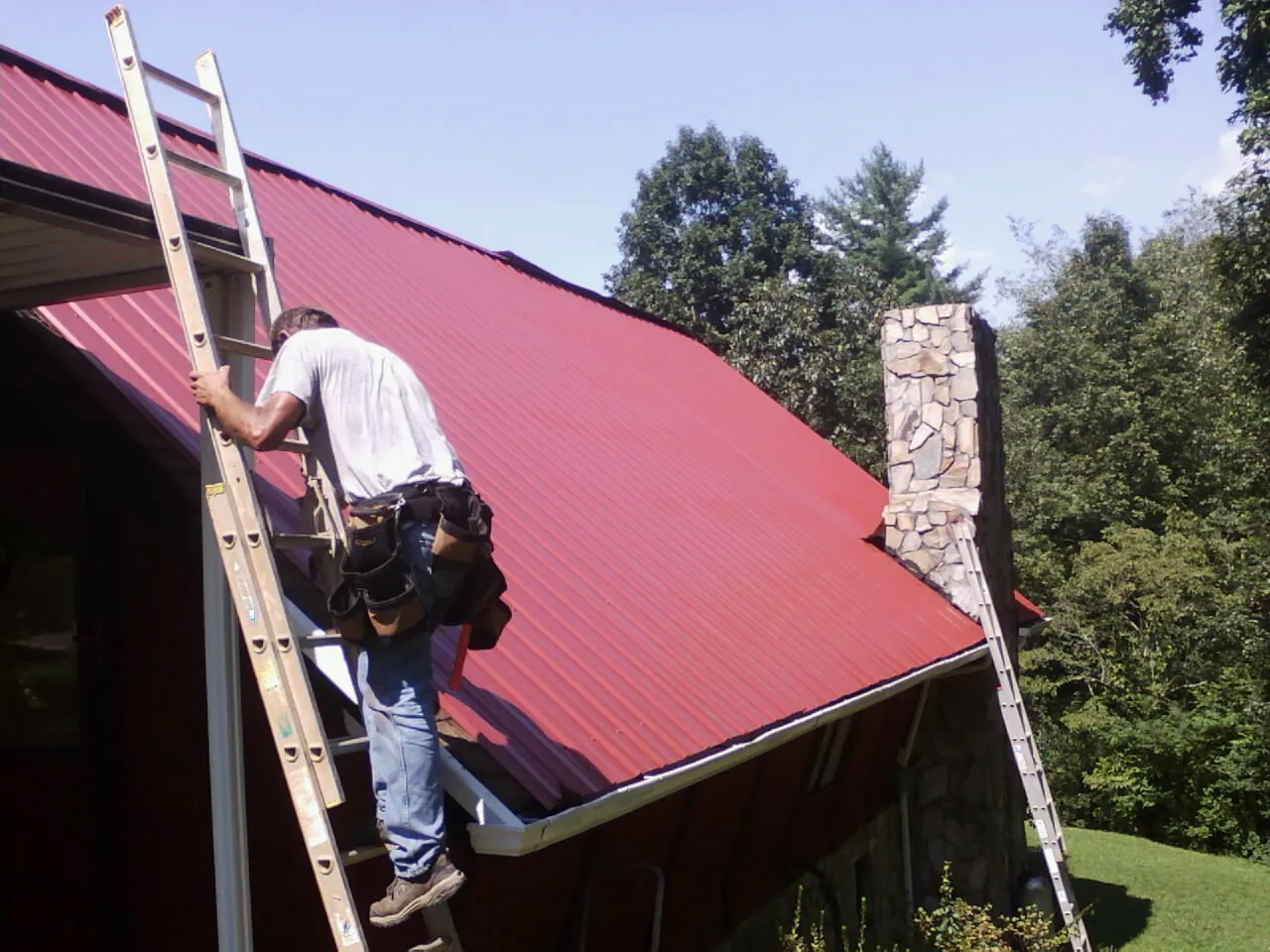 Roofer on a ladder beside a red metal roof and stone chimney — Soffit & Fascia Repair by Roofing Brothers in Hiko